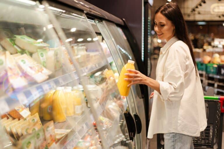 A smiling woman is lifting an orange juice container from a refrigerator shelf in a grocery store